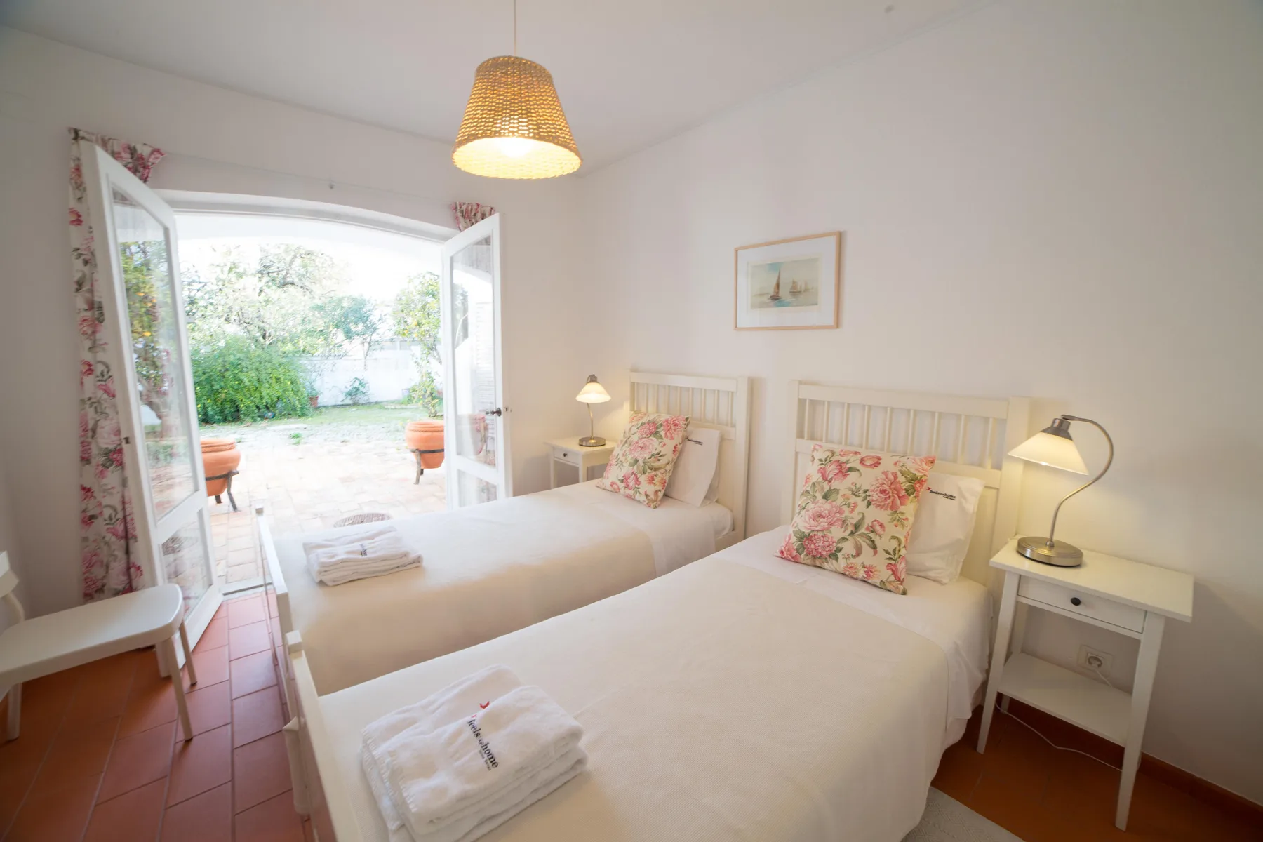 Dormitorio luminoso con dos camas, cojines florales y puertas abiertas a un patio.