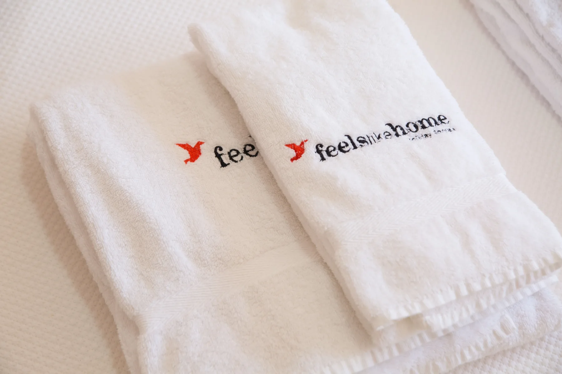 Toallas blancas con logotipos bordados están sobre una superficie clara y texturizada.