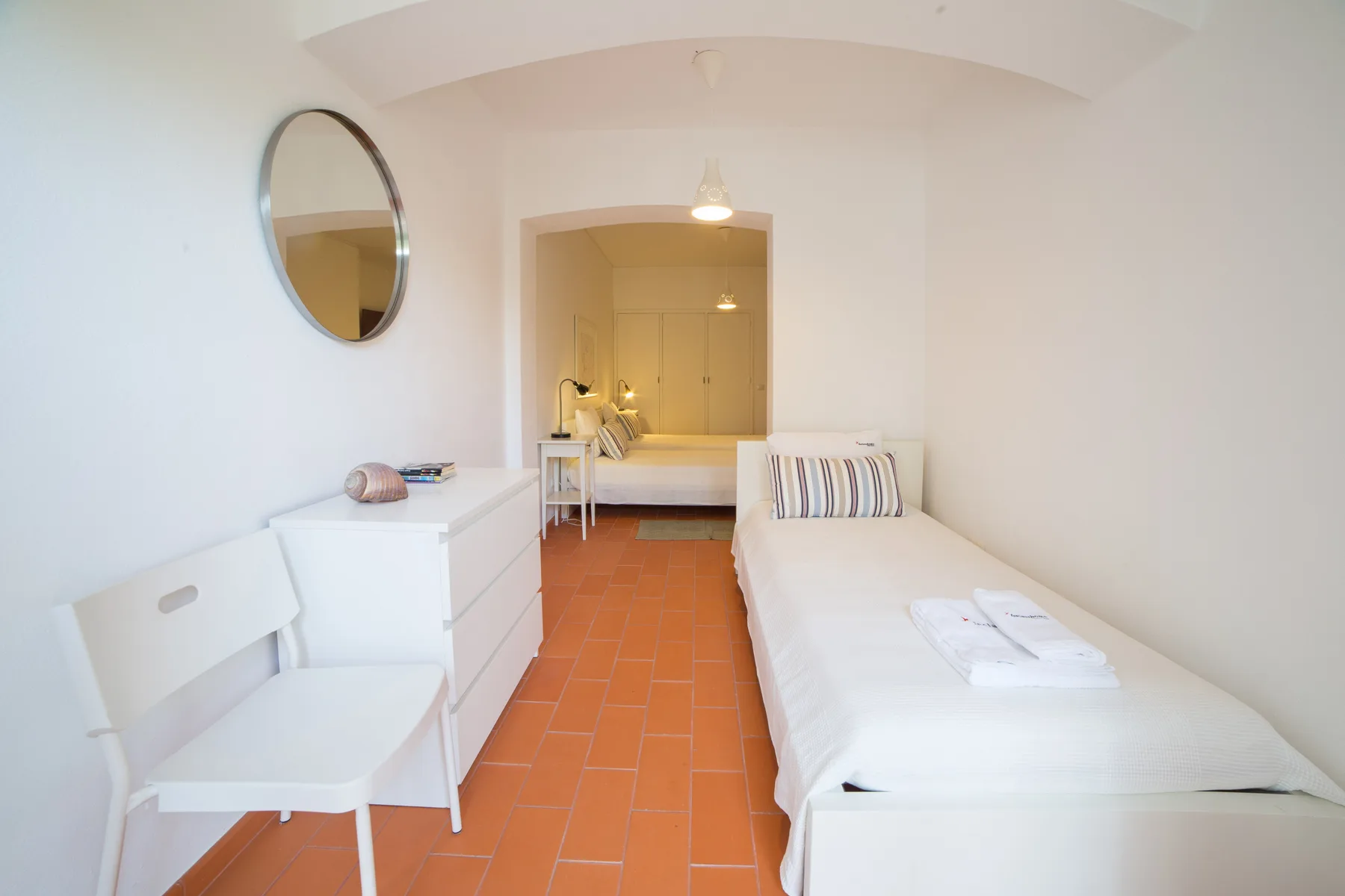 Un dormitorio estrecho con una cama individual, muebles blancos y suelo de baldosas terracota.
