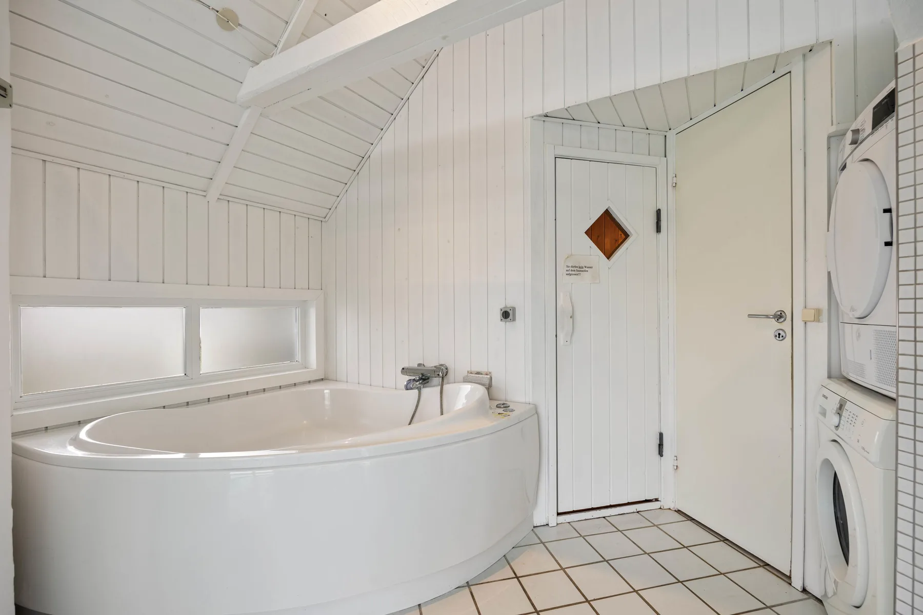 Un baño con una bañera curva, suelo de baldosas y electrodomésticos para lavar la ropa.