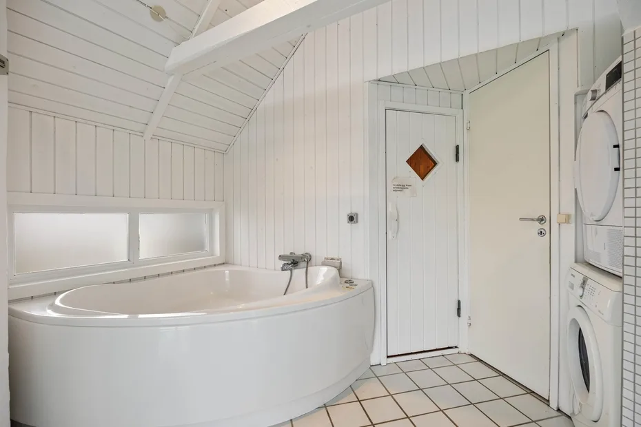 Un baño blanco con una bañera esquinera y lavadoras junto a la puerta.