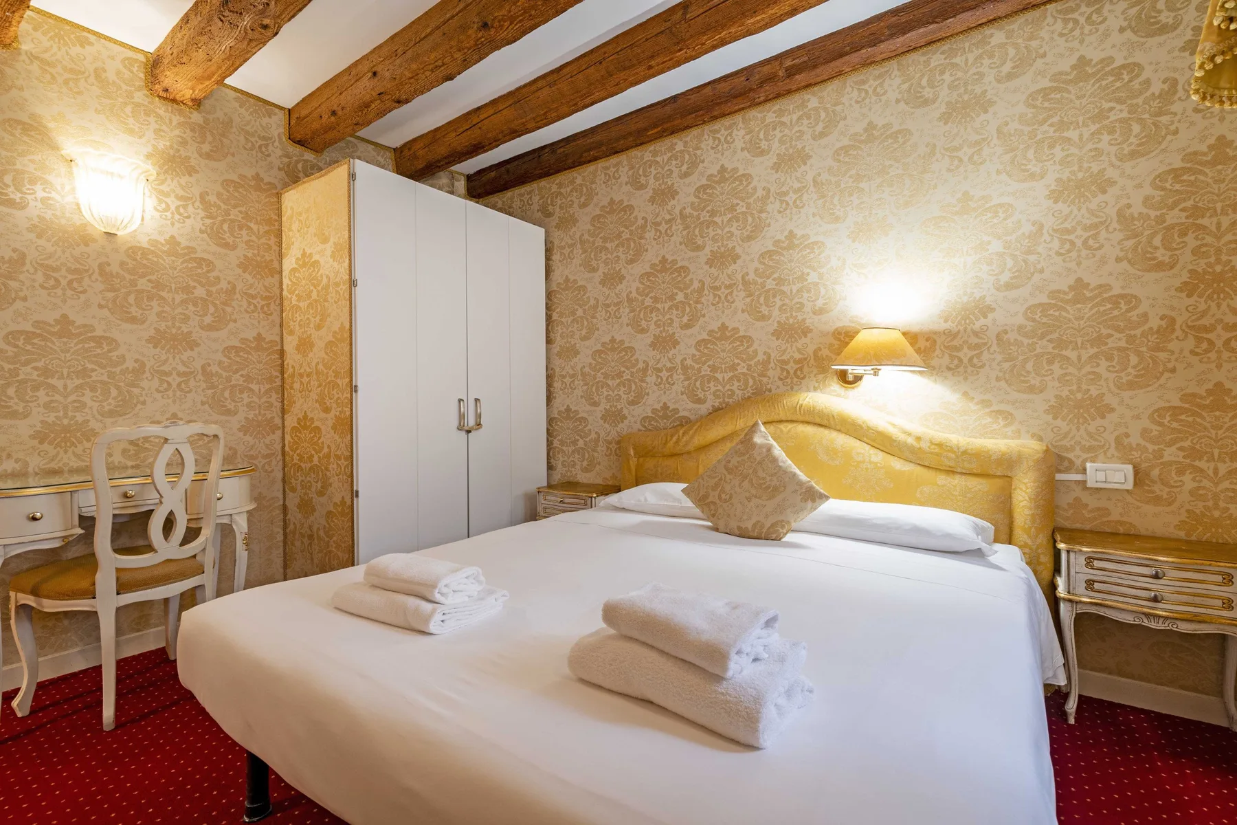 A bedroom with a double bed, patterned wallpaper, and a small writing desk.