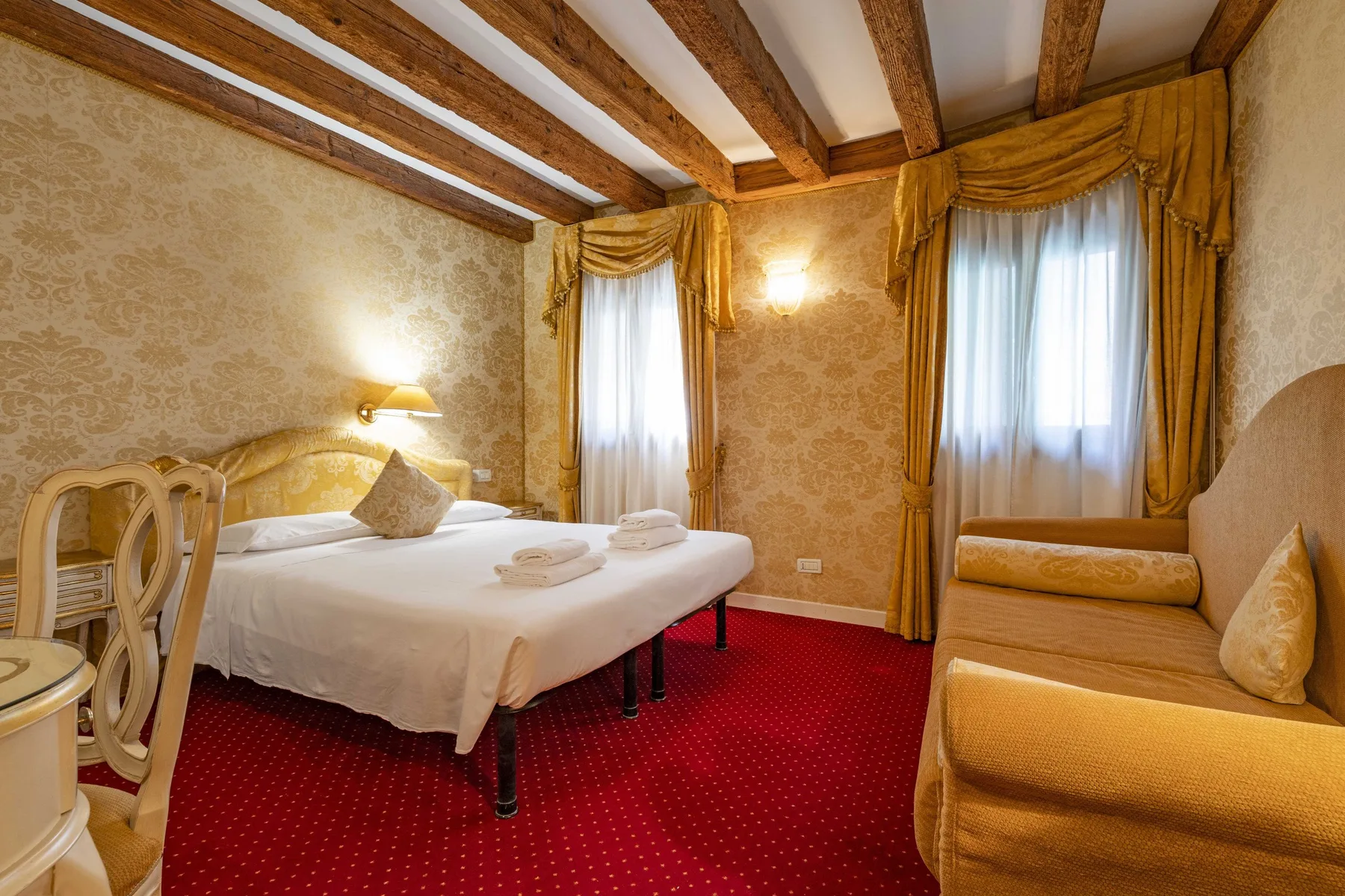 A bedroom with a bed, seating, and red carpet beneath exposed ceiling beams.