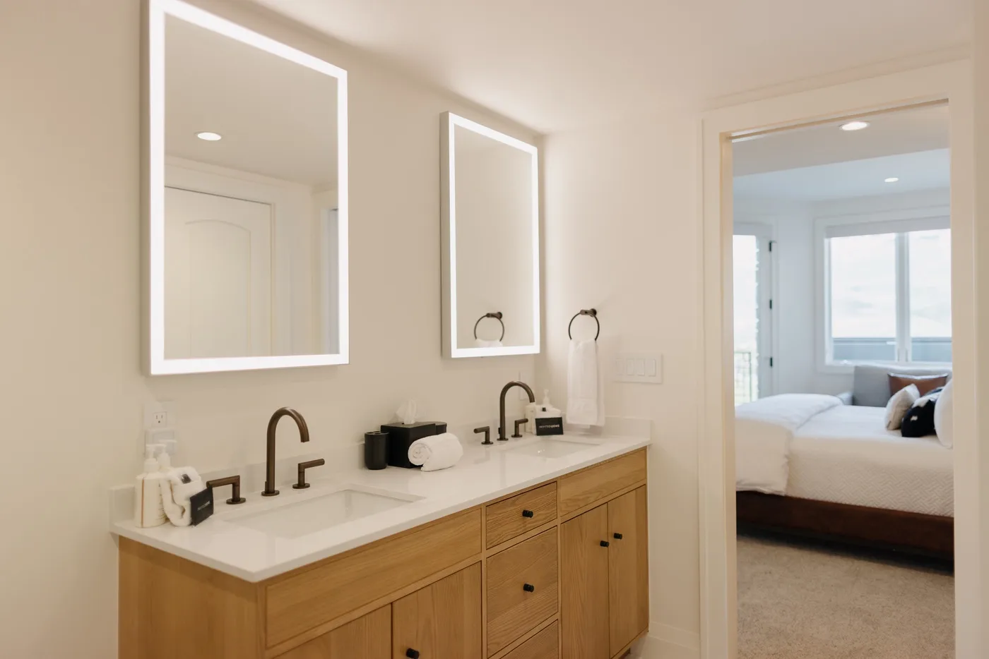A bathroom vanity with a sink and mirrors opens onto a bright bedroom.