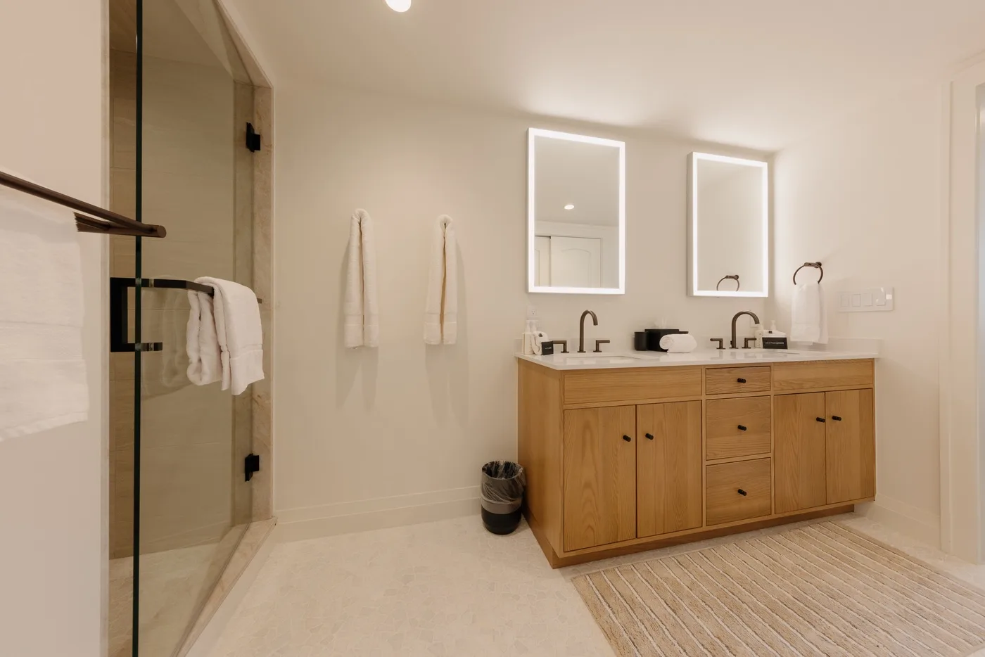 A bright bathroom with a wooden double vanity and glass shower.