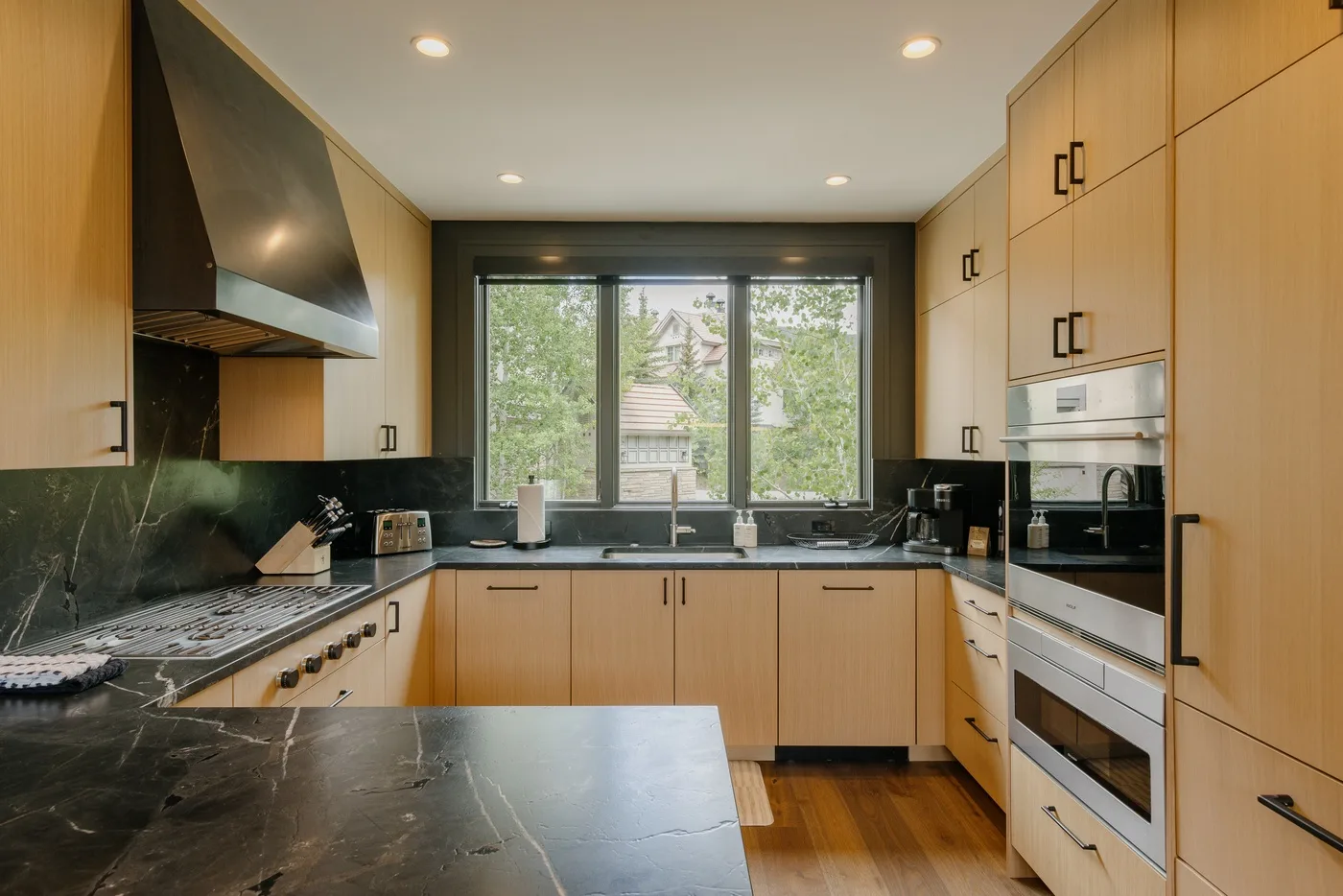 A modern kitchen with light wood cabinetry, dark countertops, and a large window.