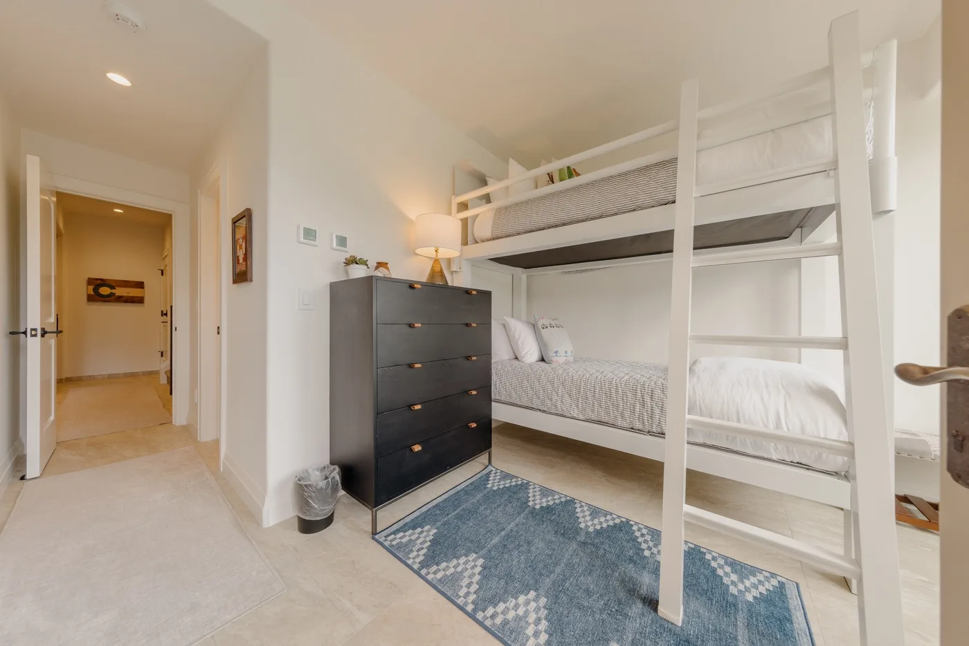 A bright bedroom with a white bunk bed, dark dresser, and blue rug.
