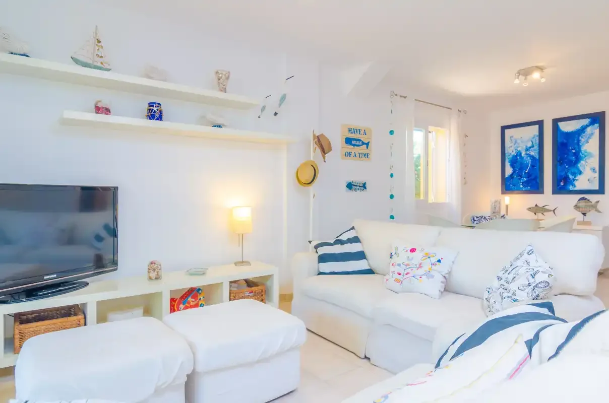 A bright living room with white seating, a television, and wall shelves.