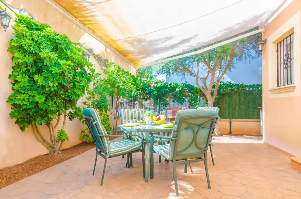 A covered outdoor dining area with a table and green seating beside leafy plants.