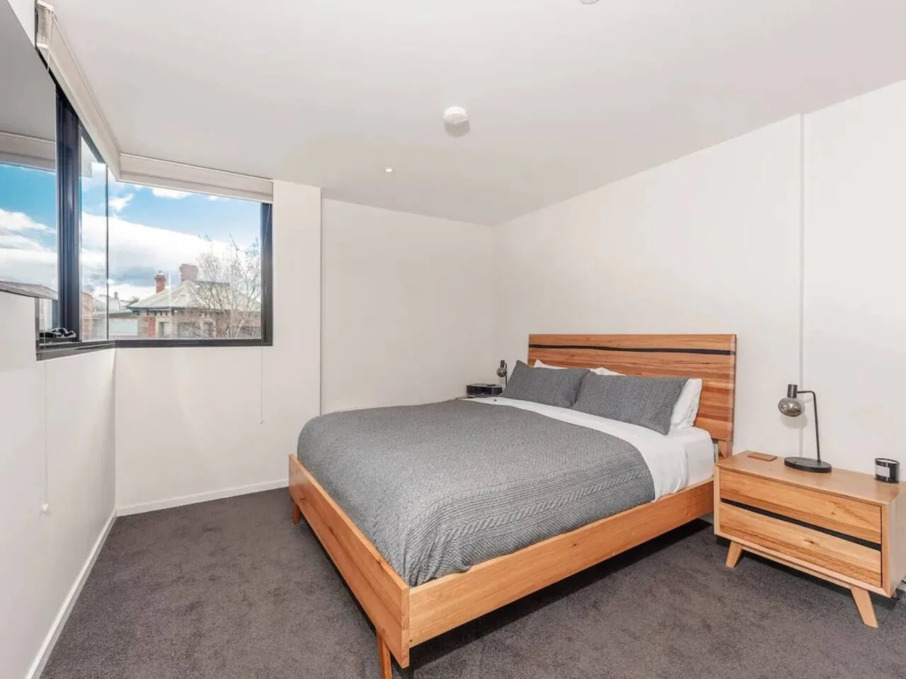 Dormitorio con una cama de madera, ropa de cama gris y una ventana amplia.