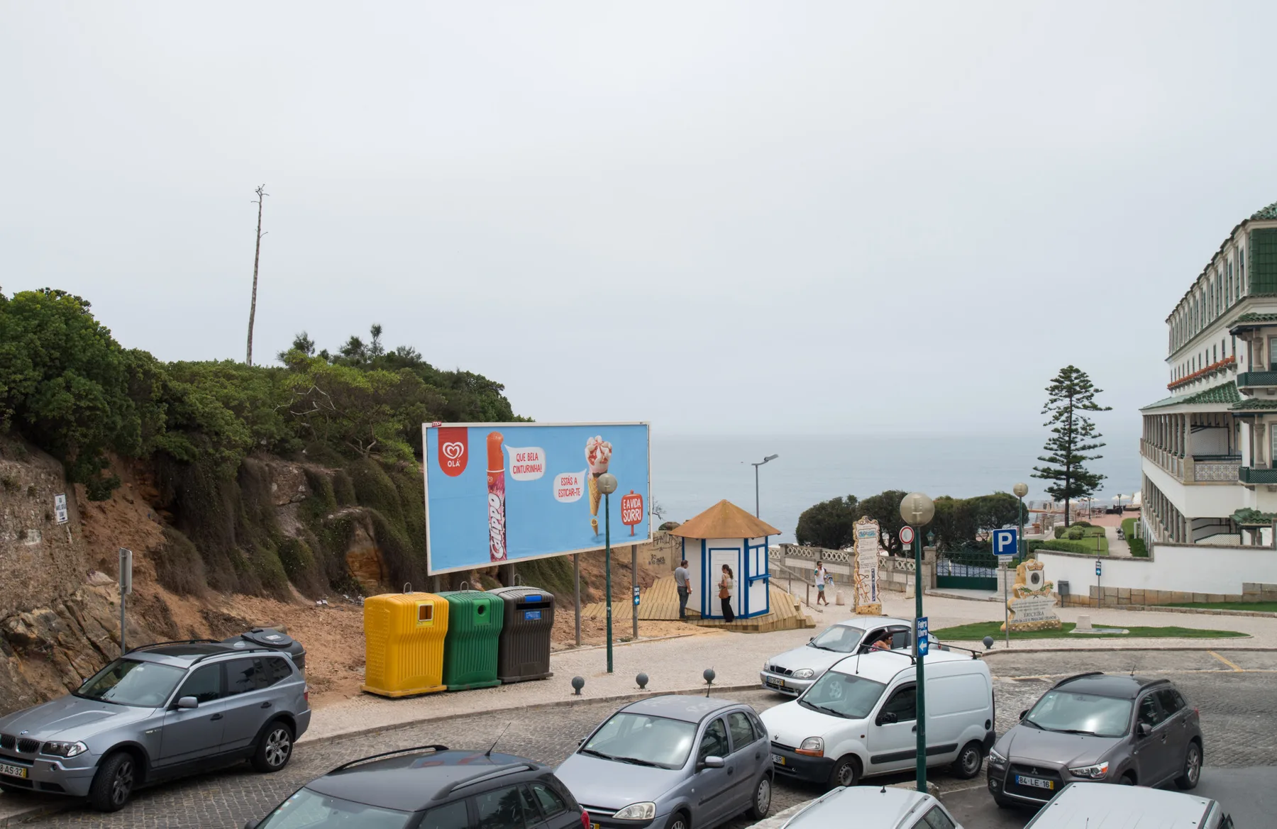 A coastal street with parked cars, a hillside, and a beachfront promenade.