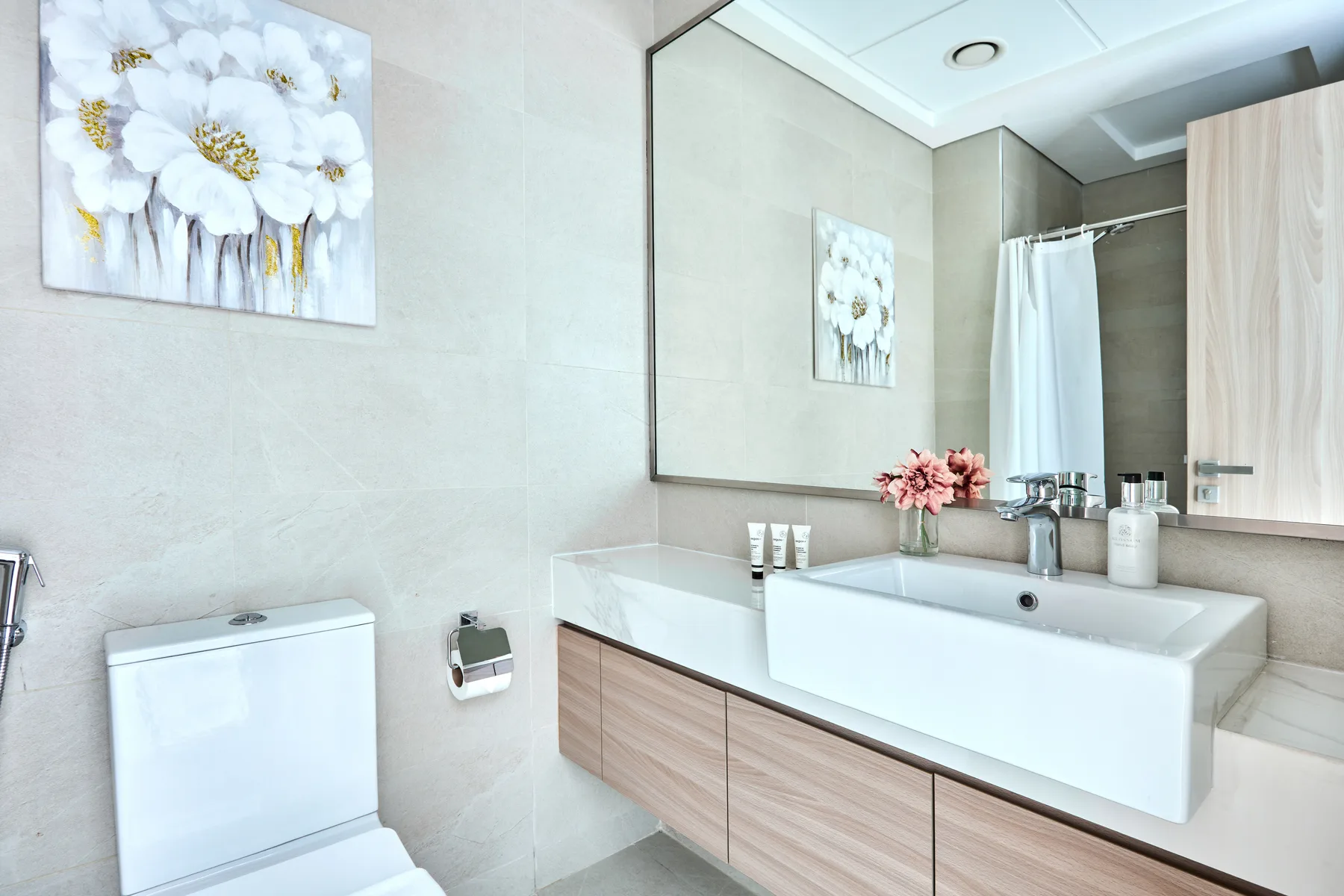 Un baño luminoso con un amplio lavabo, espejo de pared y cuadro floral.