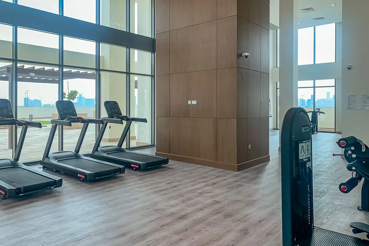 Un gimnasio luminoso con cintas de correr junto a ventanas de piso a techo.