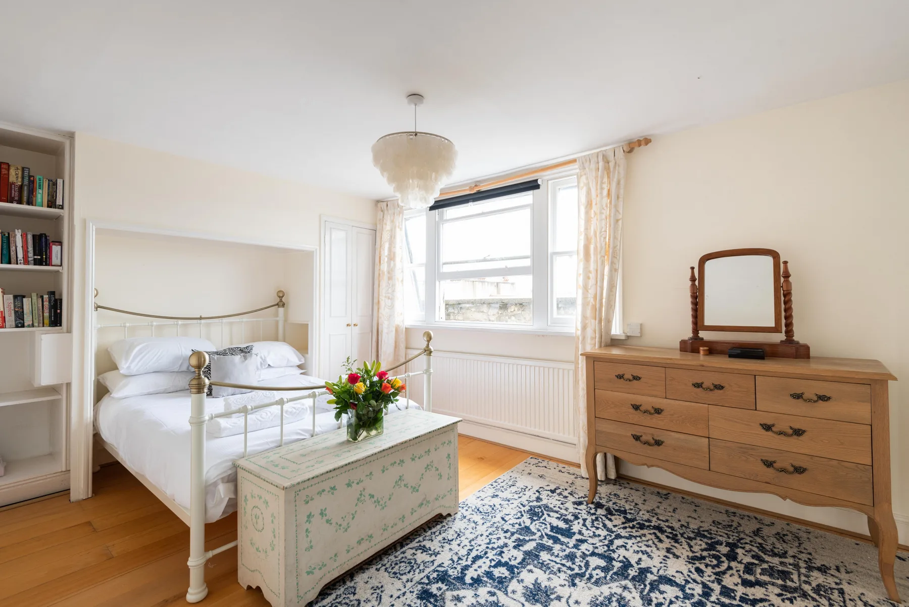 A bright bedroom with a white bed, wooden dresser, and patterned rug.