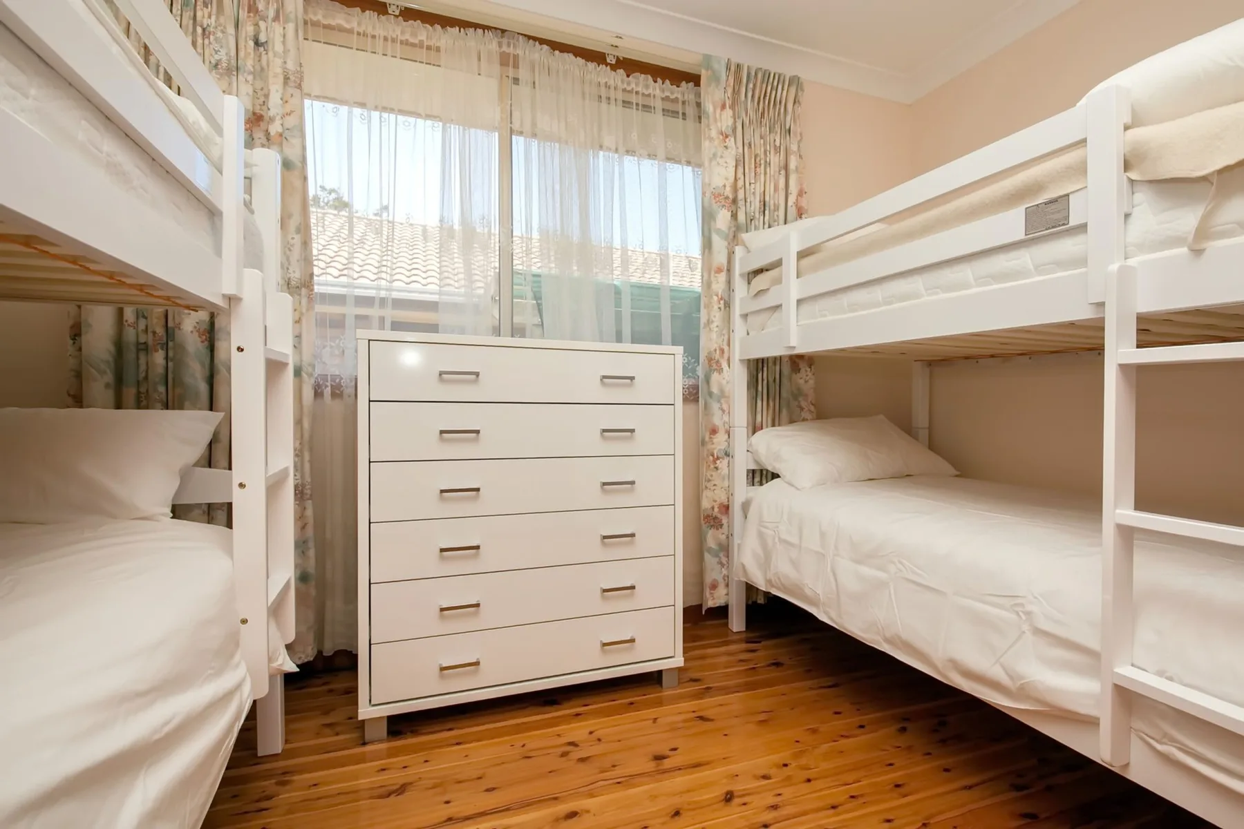 Un dormitorio luminoso con literas blancas, una cómoda y una gran ventana con cortinas.