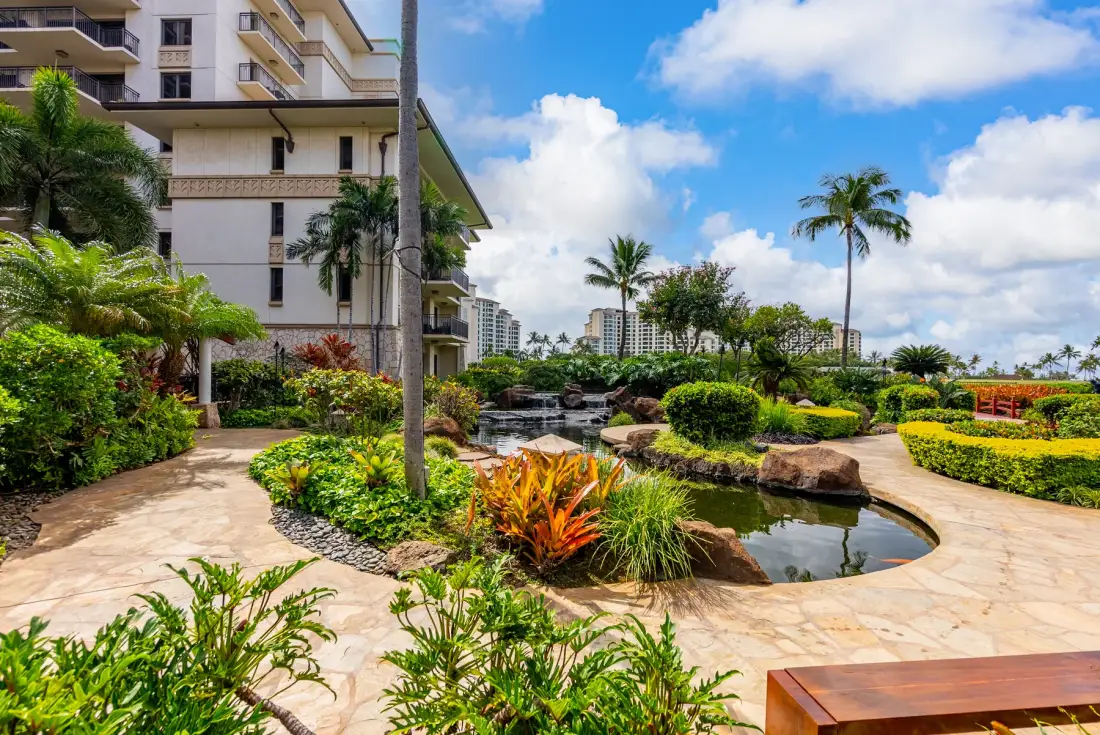 Un patio ajardinado con un estanque curvo, plantas tropicales y un camino pavimentado.