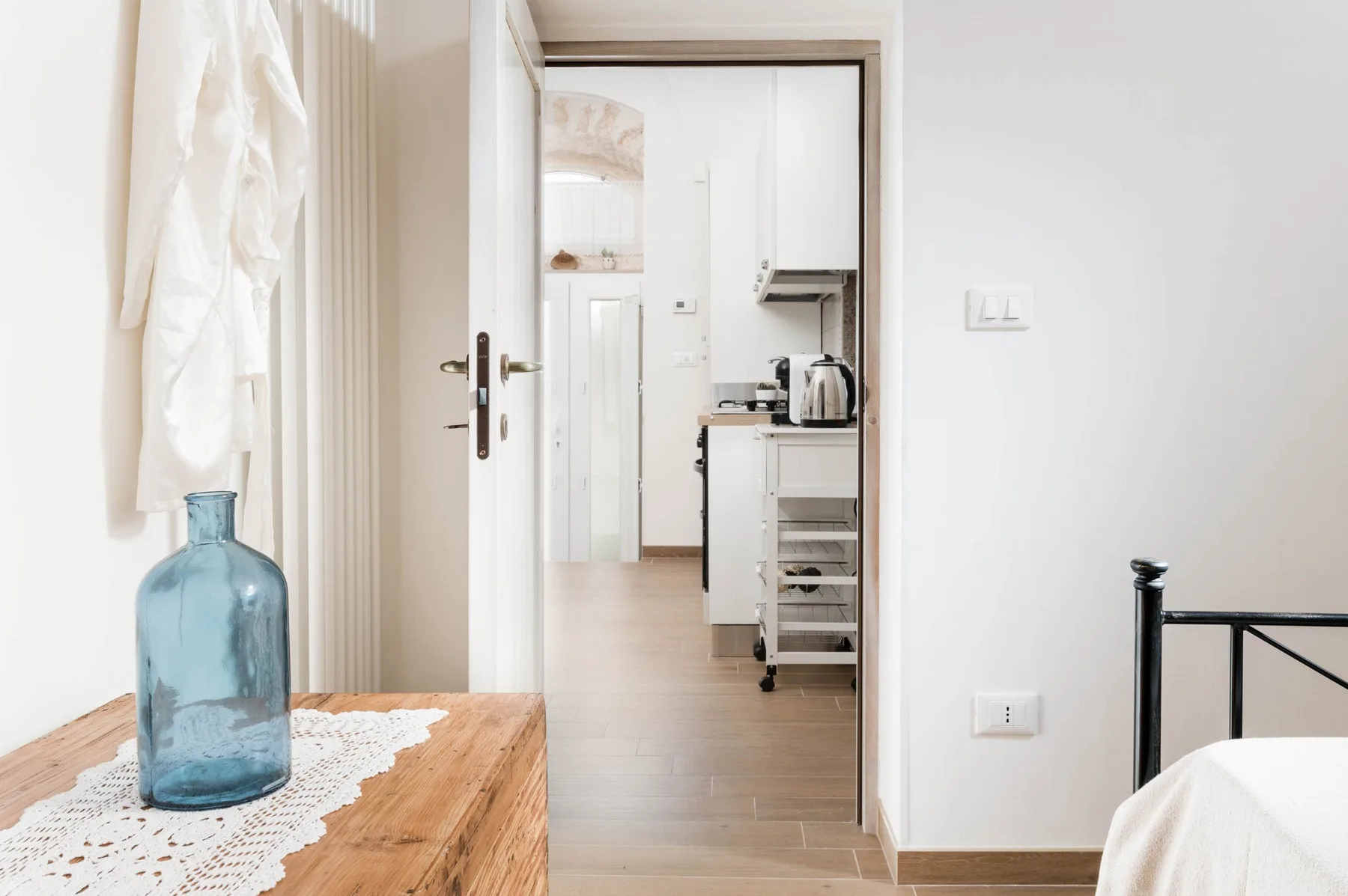 A bedroom doorway opens toward a bright kitchen, with a wooden table in the foreground.