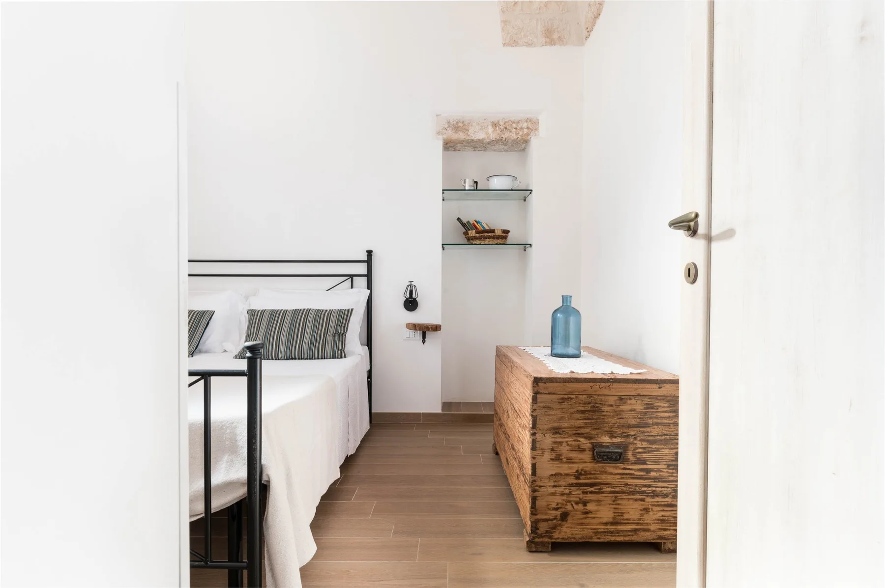 A bright bedroom with a bed, a wooden chest, and open wall shelves.