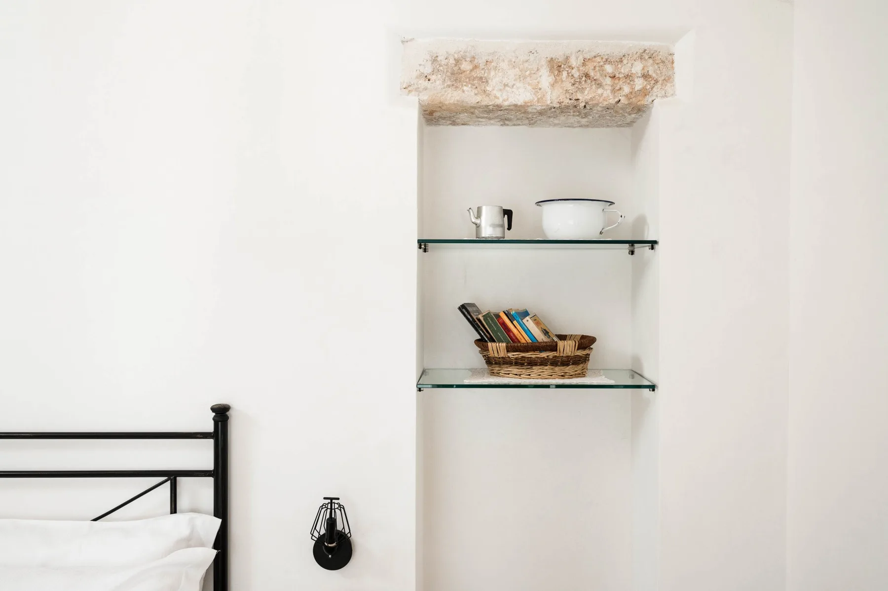 A bedroom corner with a black bed frame beside recessed shelves holding books and dishes.