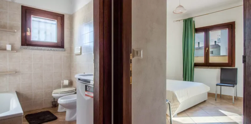 A tiled bathroom opens onto a bedroom with a bed and green curtains.