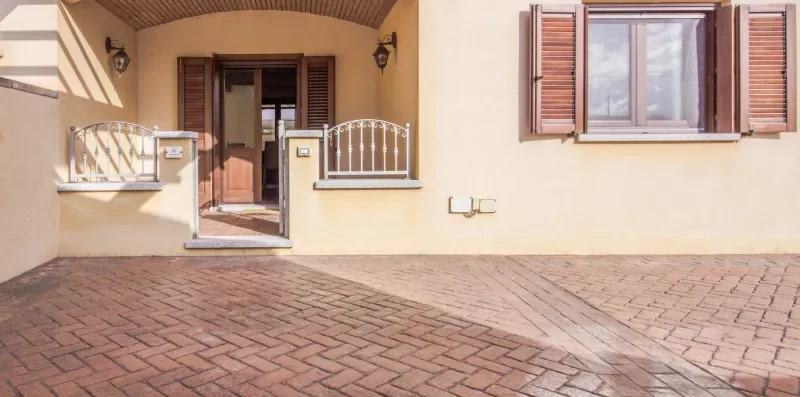 A paved patio sits in front of a beige building with a doorway and windows.