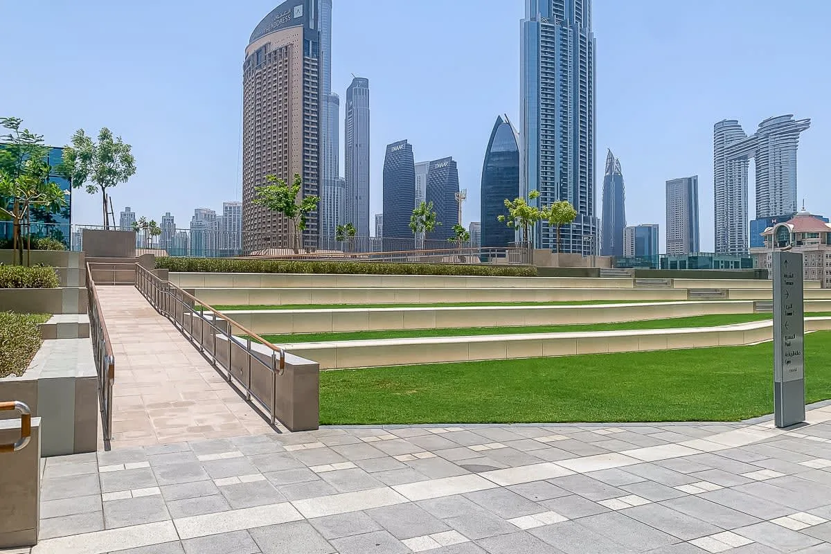 A landscaped urban plaza with a paved walkway, green lawn, and high-rise buildings.