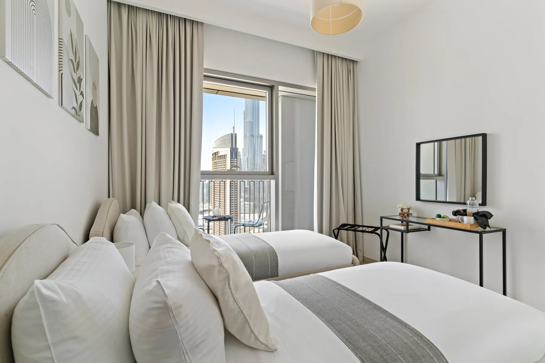 A bright bedroom with white bedding and a balcony window overlooking city buildings.