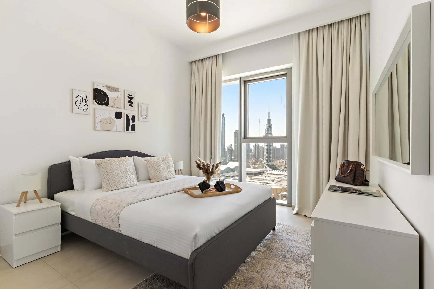 A bedroom with a bed, bedside table, and large window overlooking city buildings.