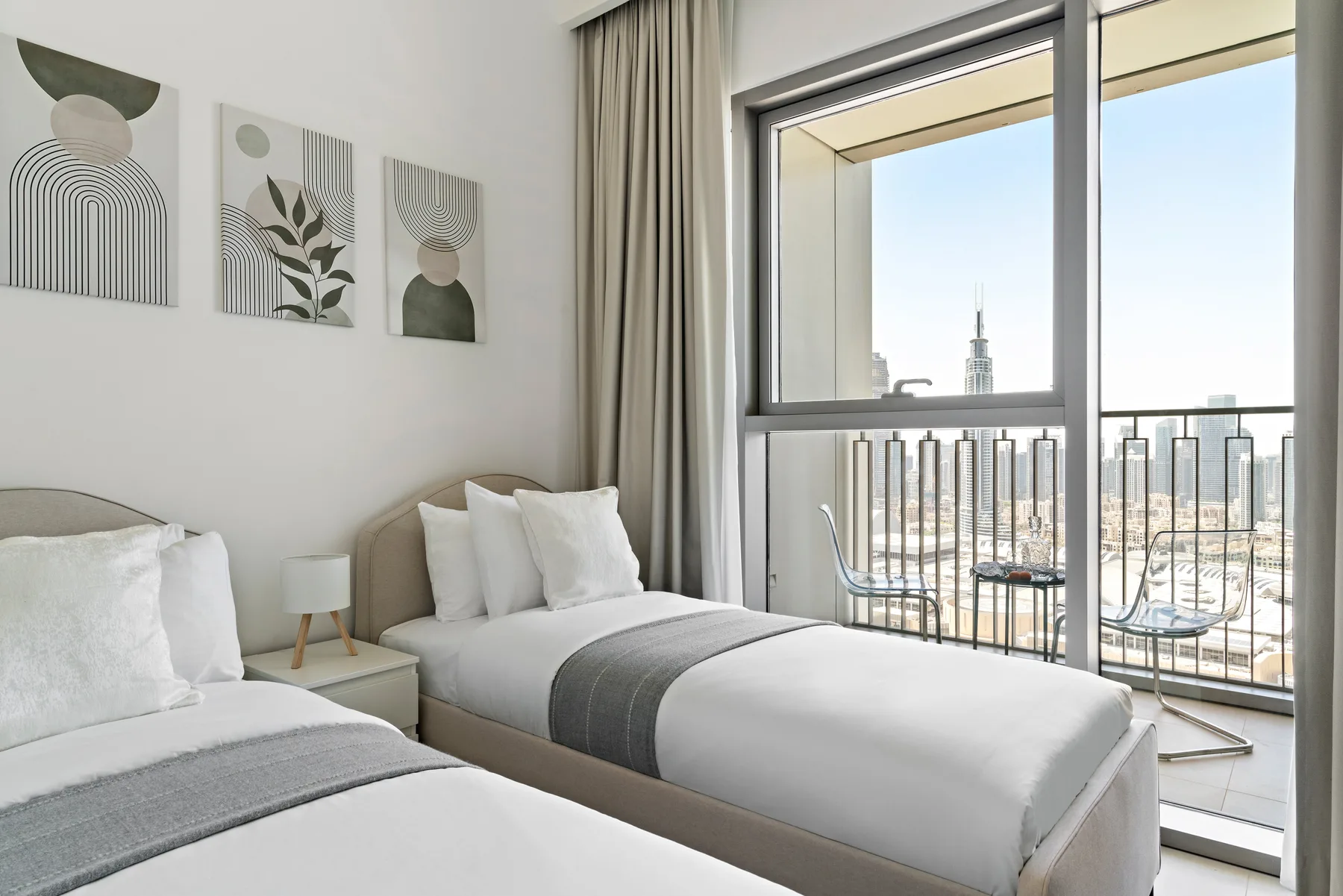 A bright bedroom with two beds beside large windows overlooking the city.
