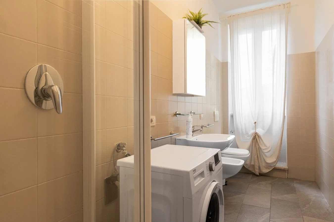 Un baño luminoso con ducha, lavabo y lavadora junto a una ventana con cortinas.
