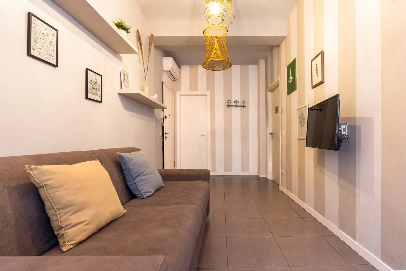Una sala estrecha con un sofá, un televisor montado en la pared y paredes a rayas.