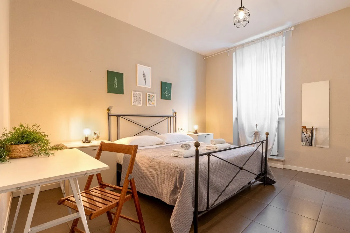 Un dormitorio con una cama de metal y una pequeña mesa junto a una ventana alta.