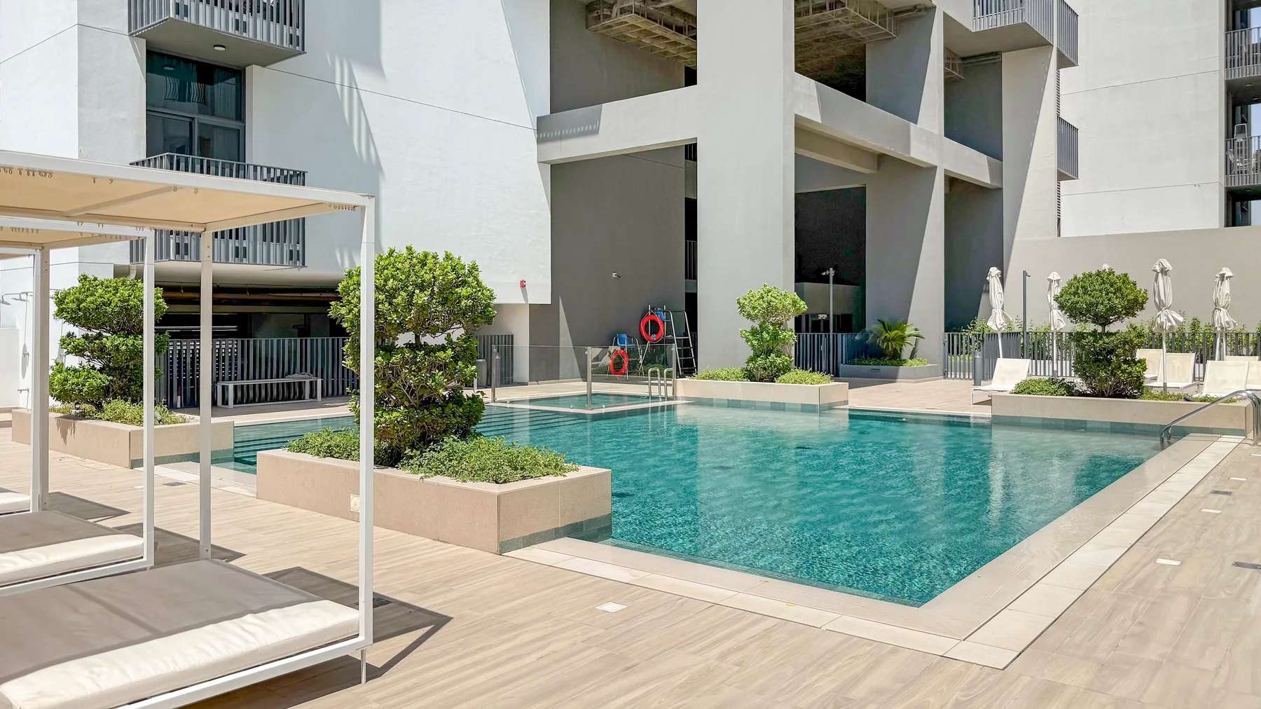 An outdoor pool beside modern buildings, with lounge seating and planted areas.