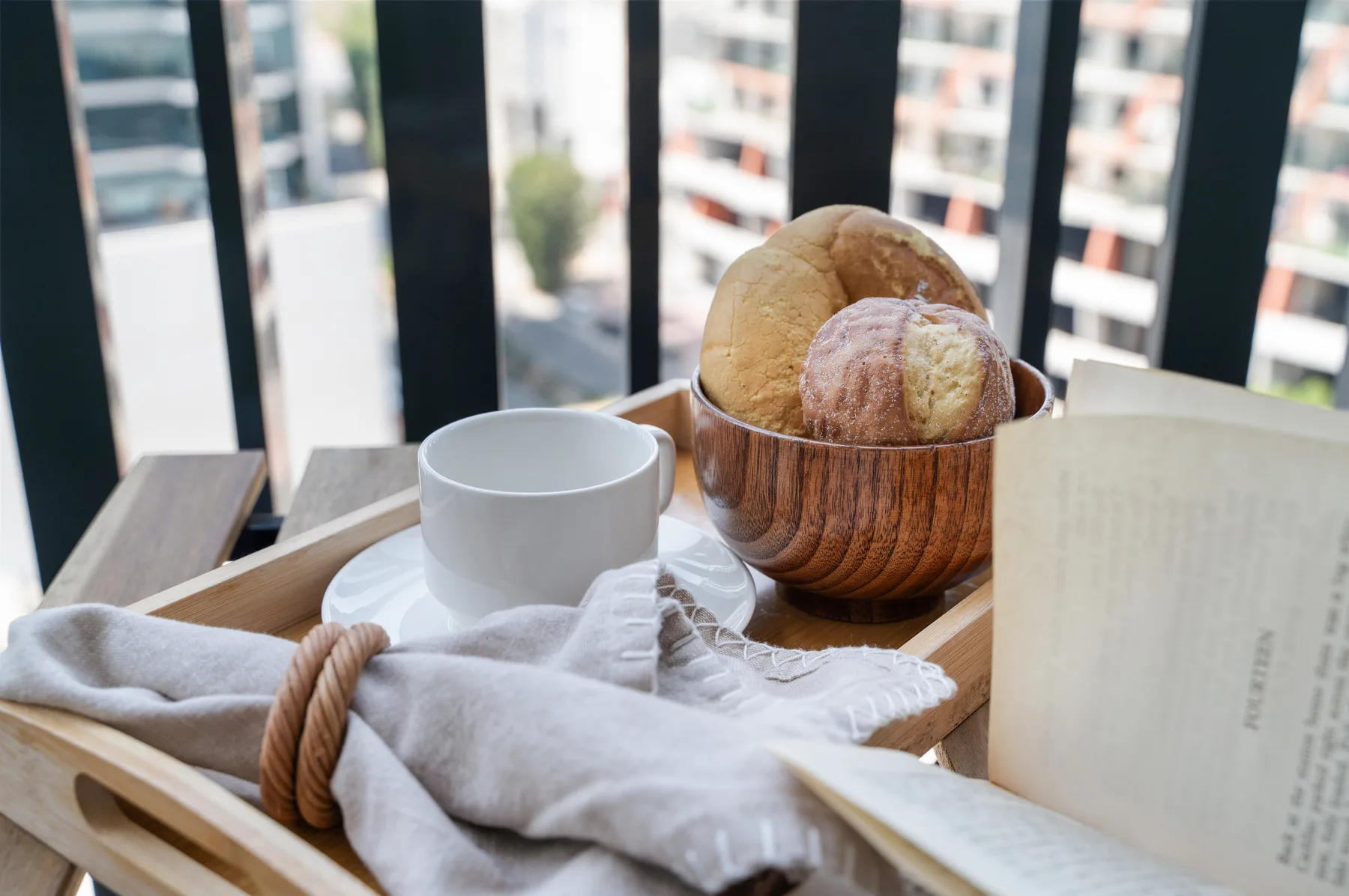 A breakfast tray by a window holds a cup, bread, and a folded cloth.