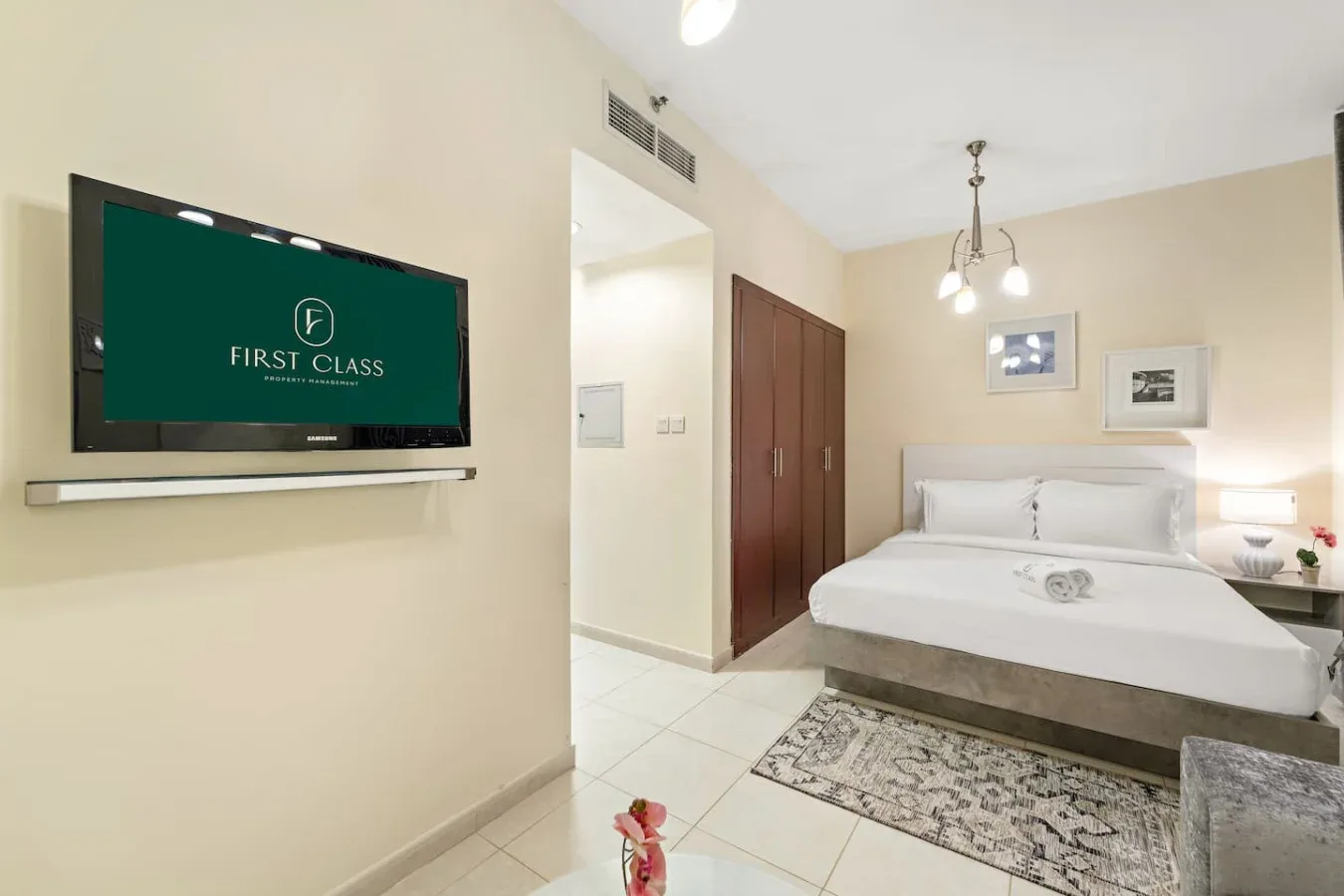 Un dormitorio luminoso con una cama bien hecha, televisor montado en la pared y alfombra estampada.