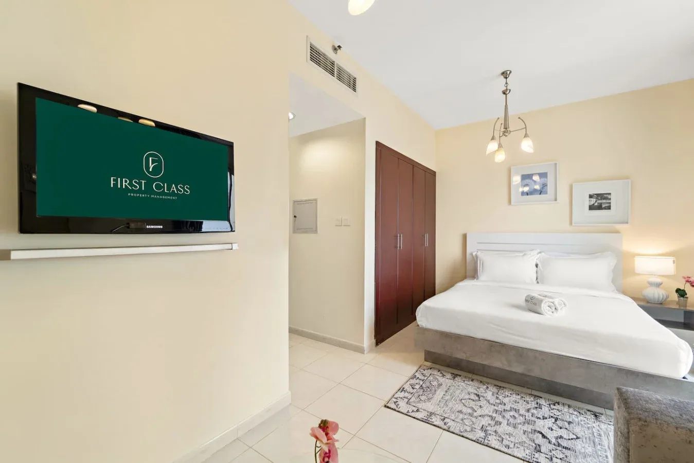Un dormitorio con una cama bien tendida, un televisor montado en la pared y suelo de baldosas.