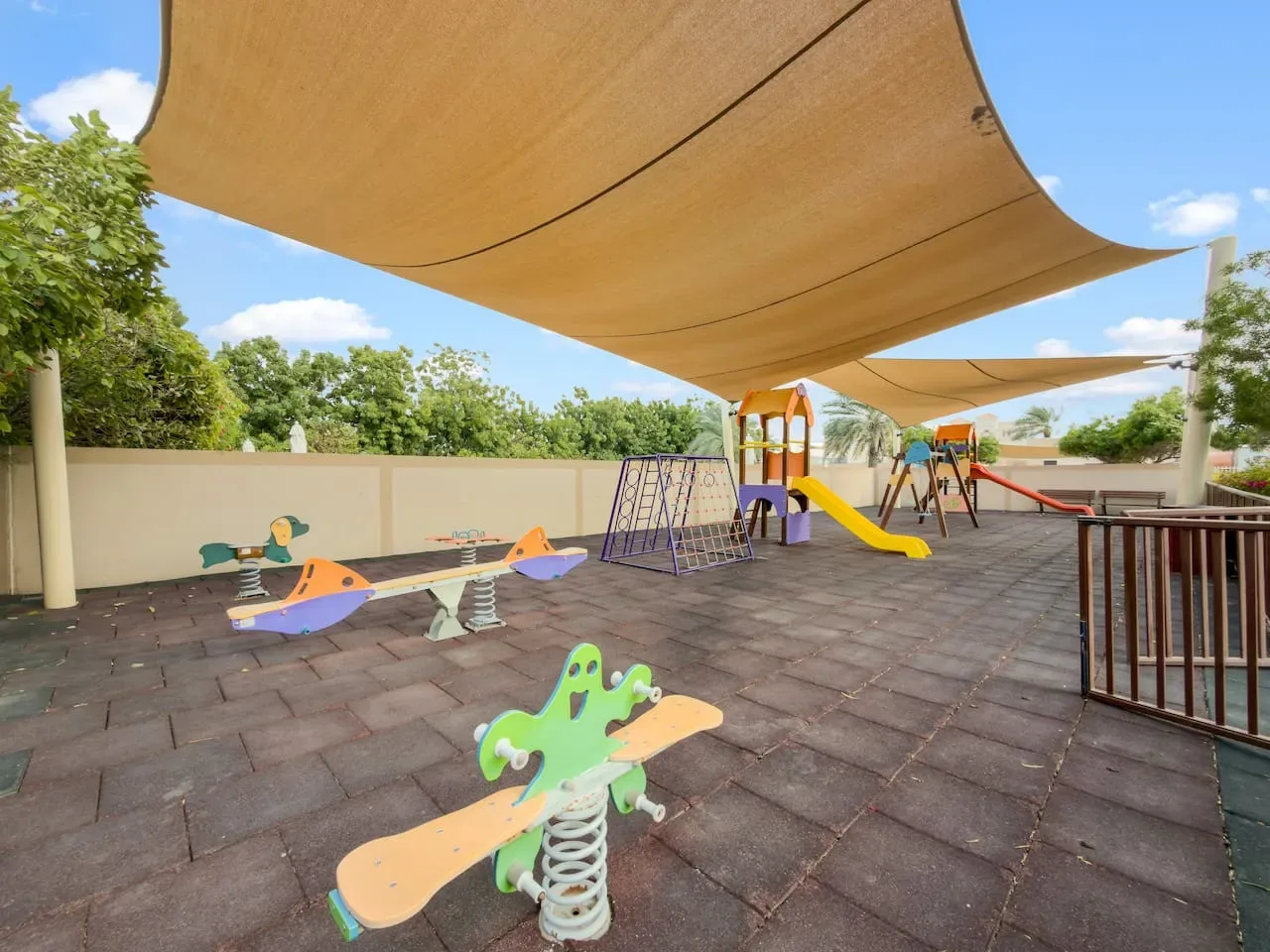 Un parque infantil al aire libre con juegos de resorte, columpios y toboganes bajo una sombra.