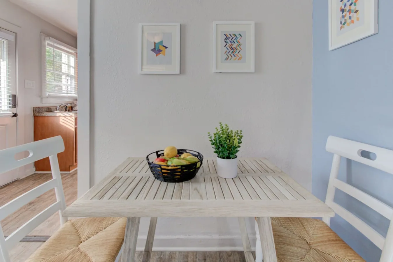 Un comedor con una mesa blanca, asientos tejidos y cuadros enmarcados.