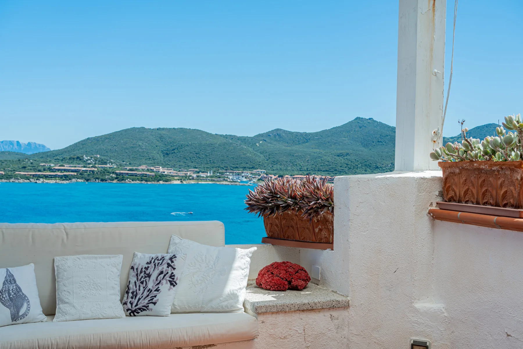 A seaside terrace with cushioned seating overlooks blue water and green hills.