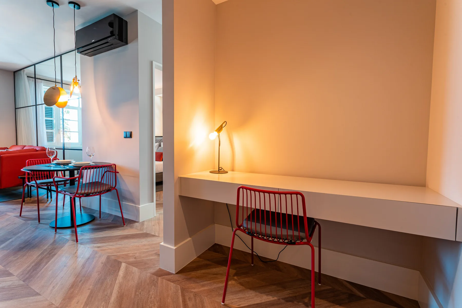 A dining area and desk nook with red seating and a lit table lamp.