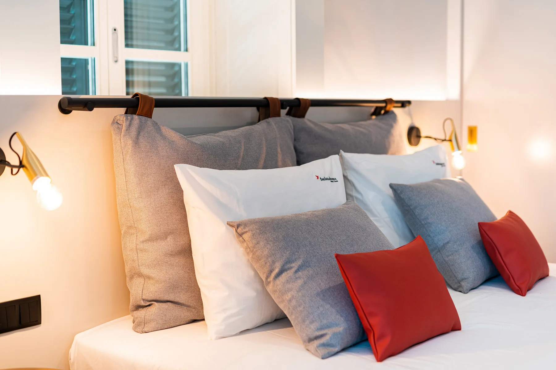 A bed with gray pillows and red cushions sits beneath a window.