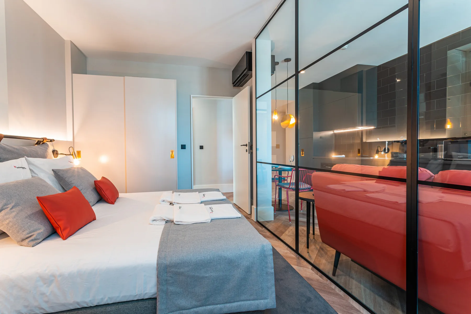 A bedroom with white bedding beside a glass partition and red seating.