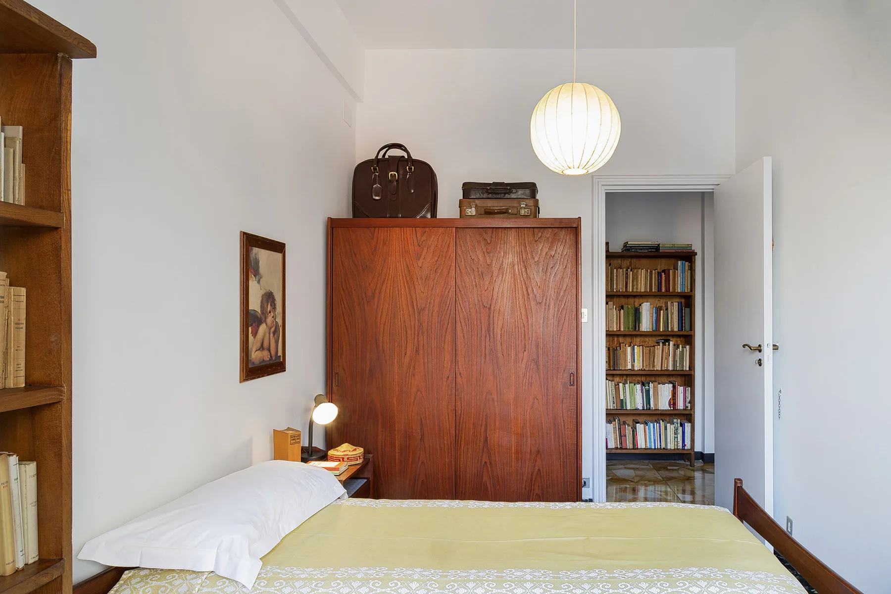 Un dormitorio con una cama, un armario de madera y estanterías visibles por la puerta abierta.