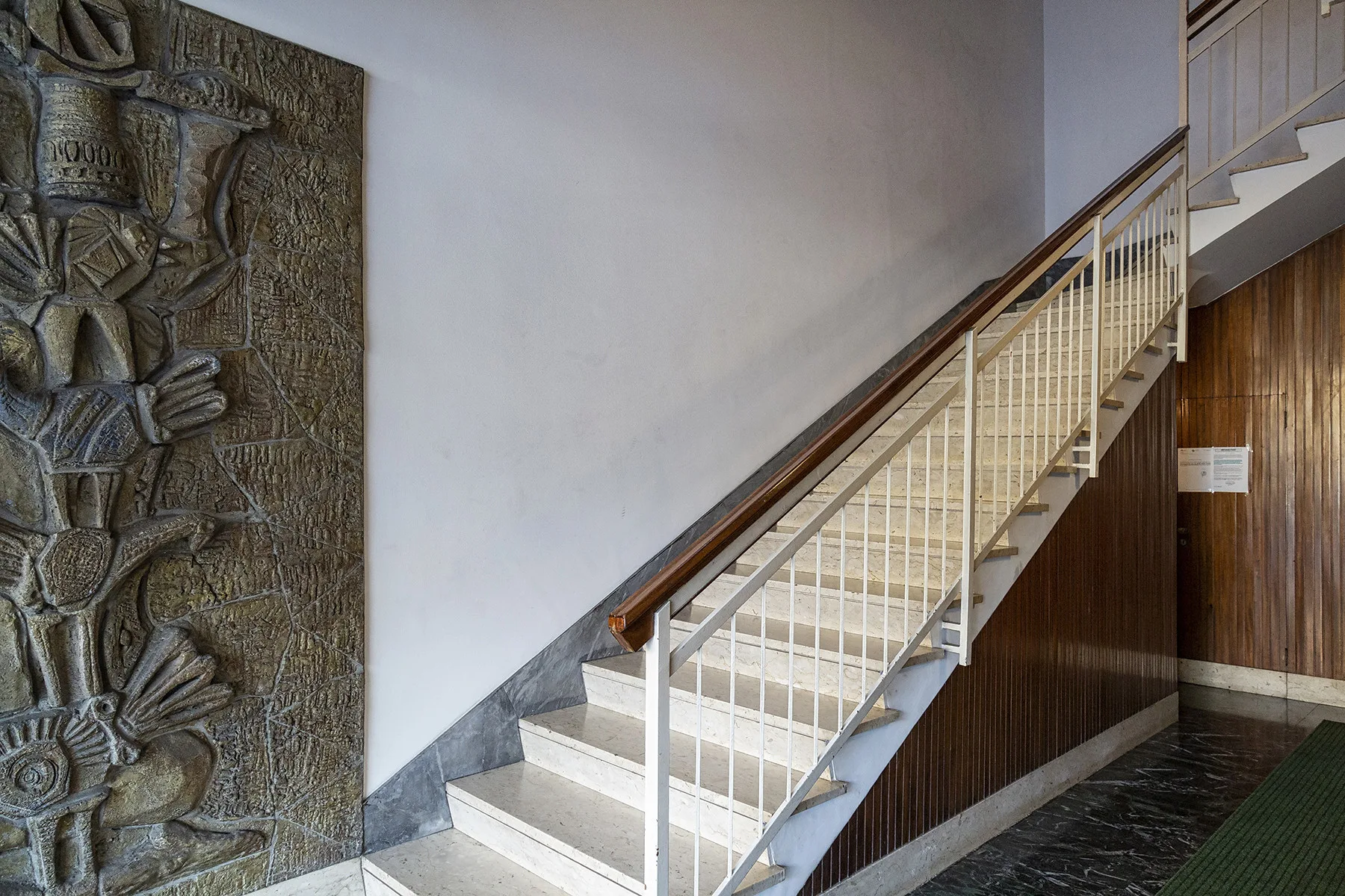 Una escalera interior con peldaños claros y barandilla metálica junto a un panel mural tallado.