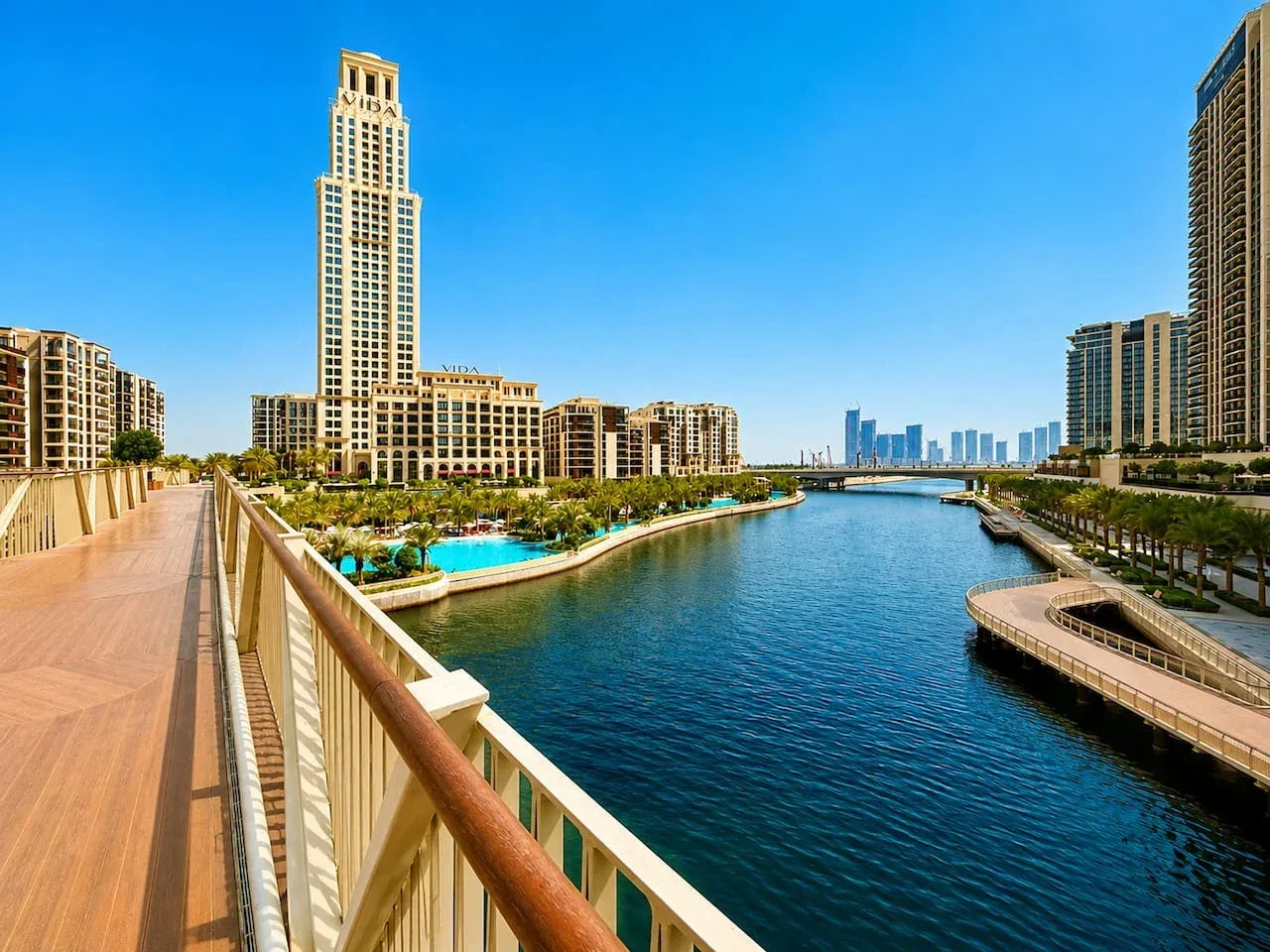Un paseo junto al agua ofrece vistas a un canal, jardines y edificios altos.