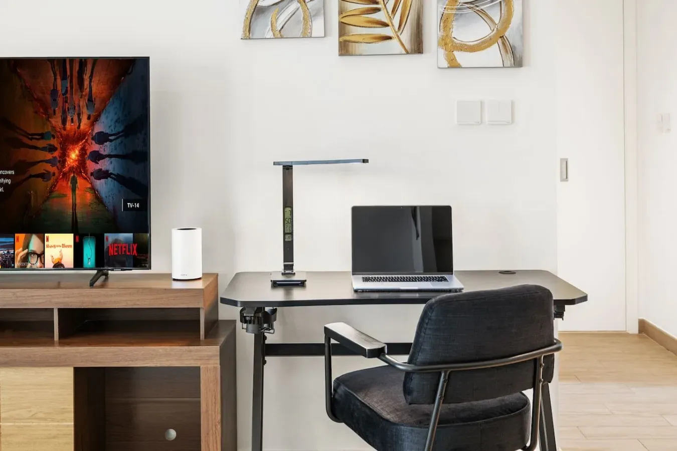 Un escritorio con portátil y silla está junto a un televisor en una habitación luminosa.