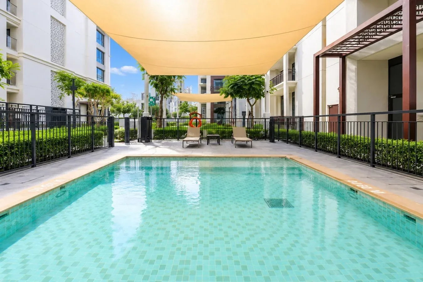 Una piscina al aire libre está bajo una amplia cubierta de sombra en un patio ajardinado.