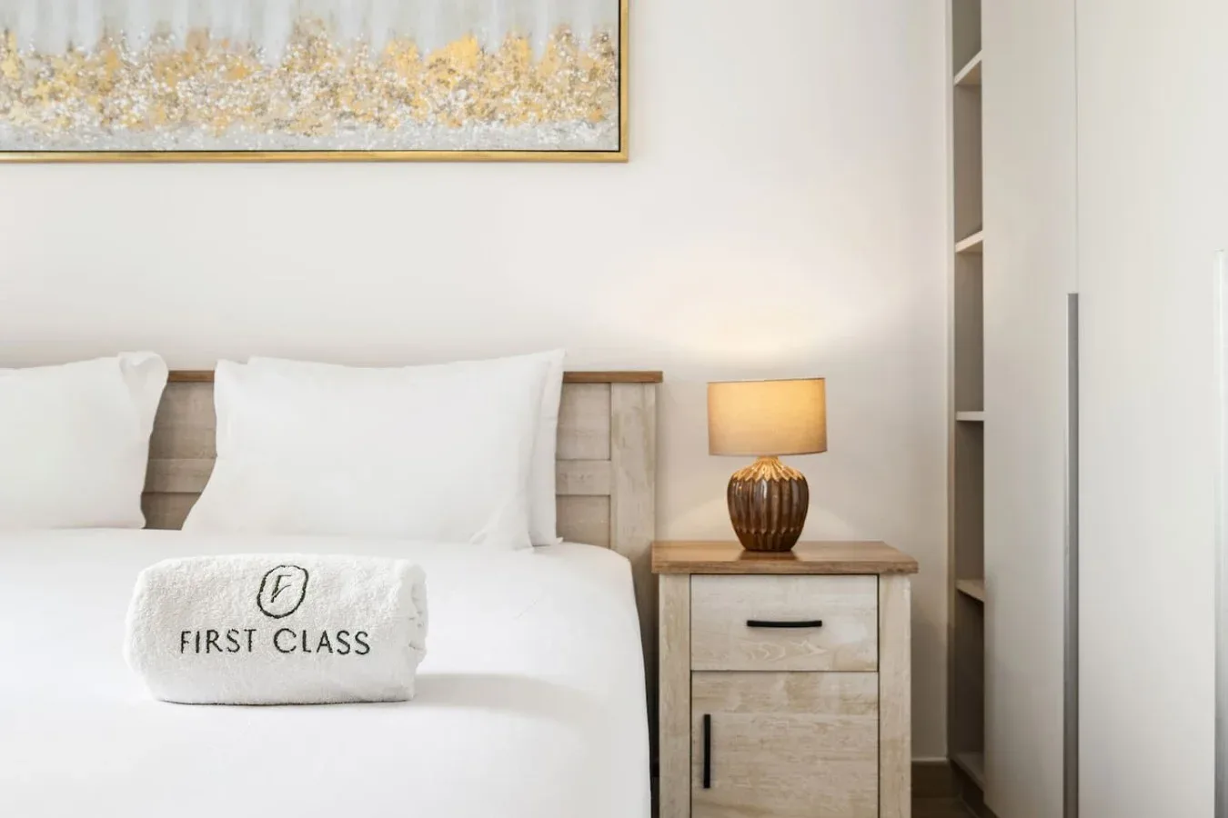 Un dormitorio con ropa de cama blanca, una mesita de noche y una lámpara encendida.