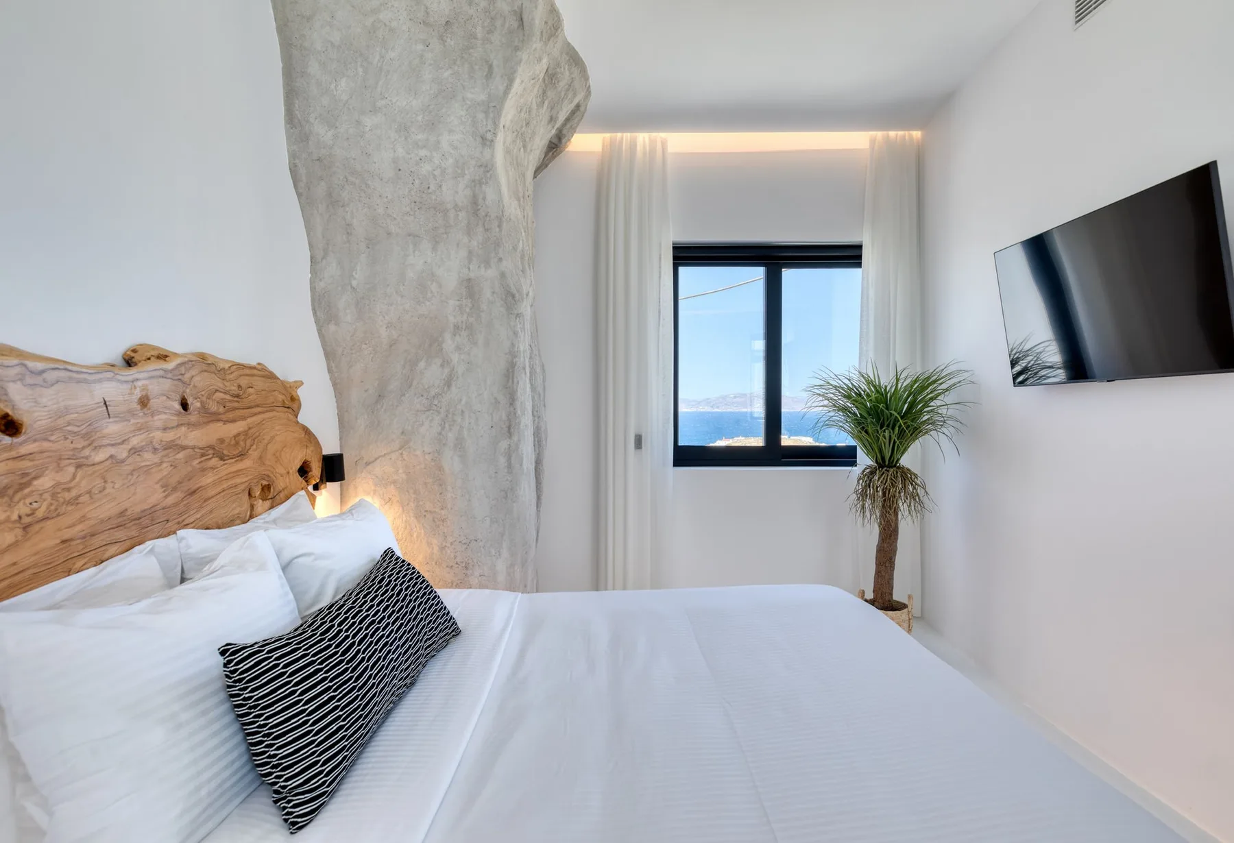Un dormitorio luminoso con ropa de cama blanca, un televisor en la pared y una ventana con vistas al mar.