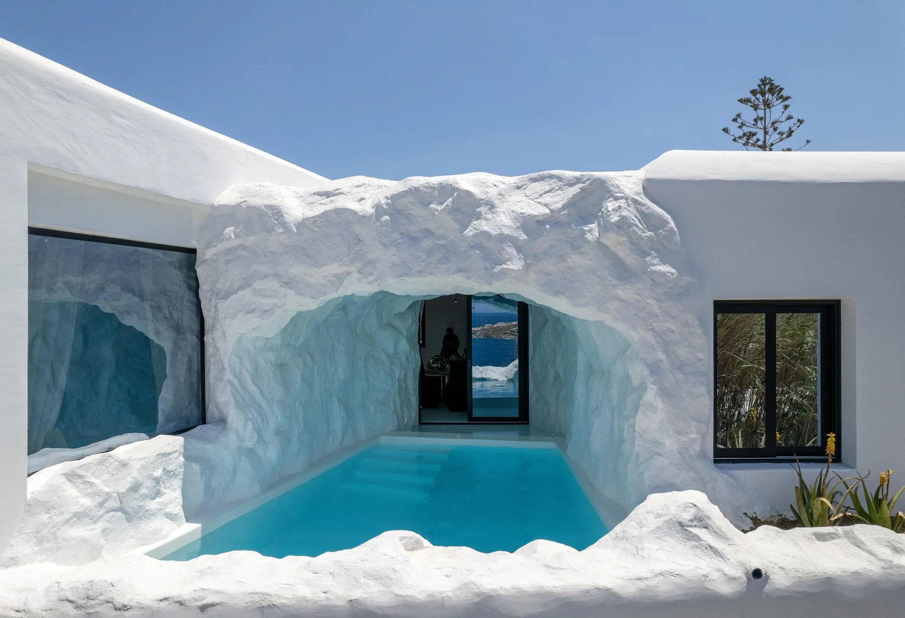A turquoise pool sits within sculpted white walls beside dark-framed windows.